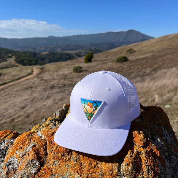 white trucker hat with poppy CA love patch sitting on a rock with mountain in the back ground