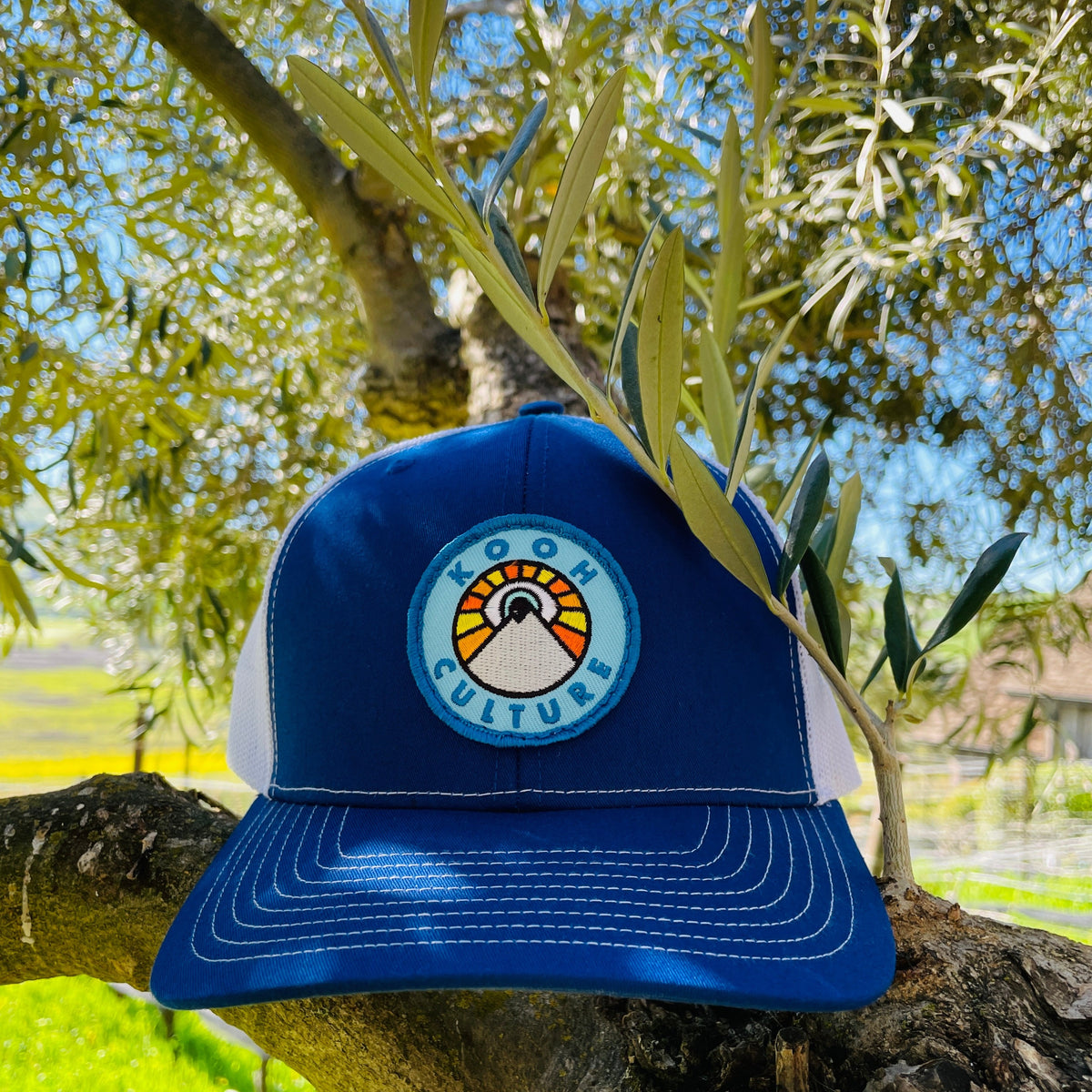 Royal blue and white Kooh Culture trucker hat with the mountain and sun badge